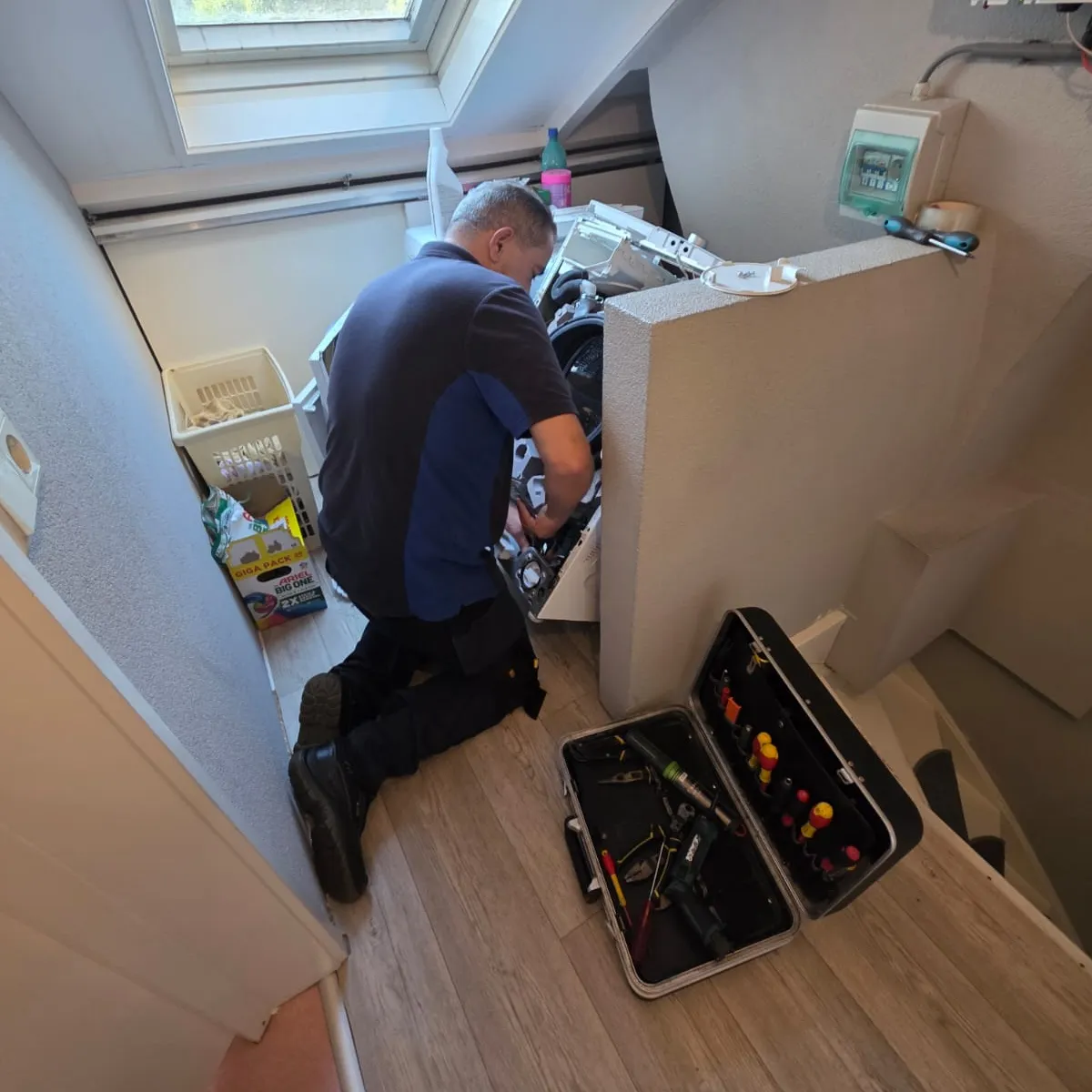 Simo geknield aan een opengewerkte wasmachine op een zolder, met zijn gereedschapskoffer open op de vloer